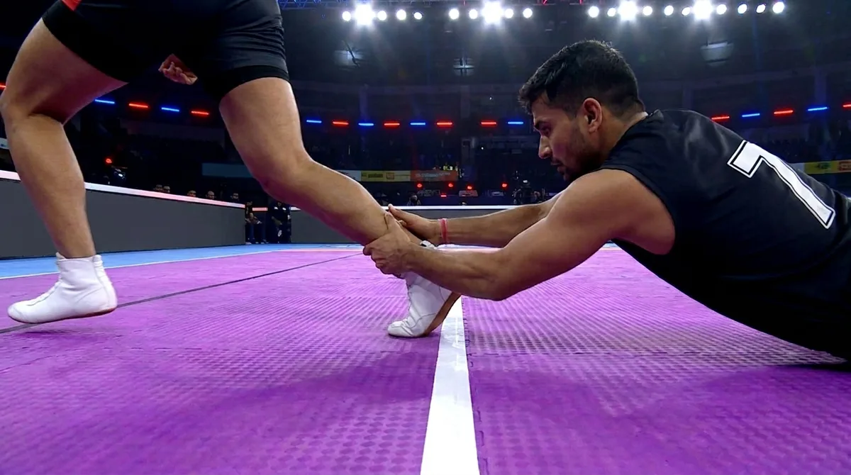 A defender stretches full length on a purple kabaddi mat to hold a raider by the ankle