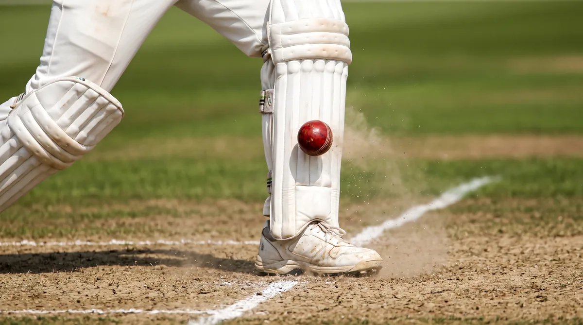 Close-up of a red cricket ball striking a batters front pad just in front of the popping crease, dust rising off the pitch