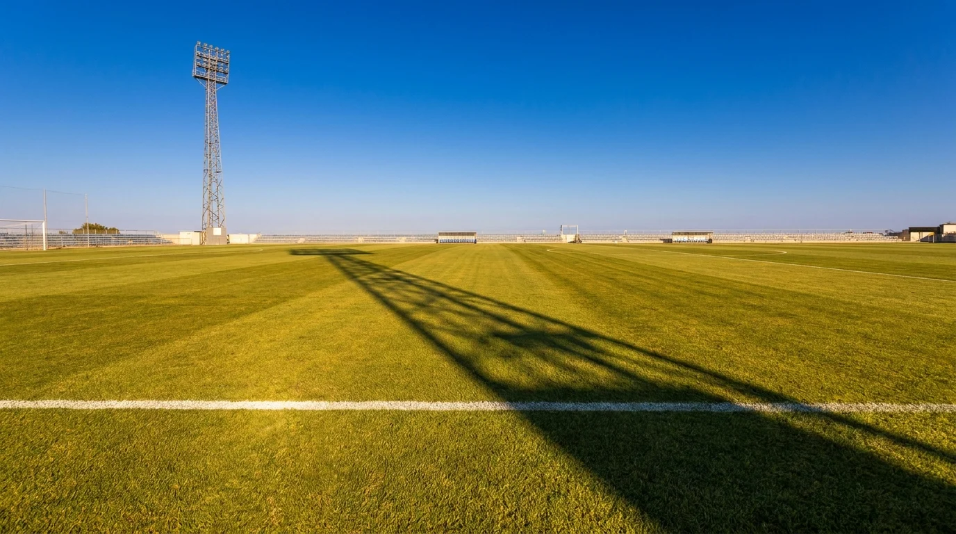 The long shadow of a floodlight pylon stretching across an empty sunlit football pitch at golden hour