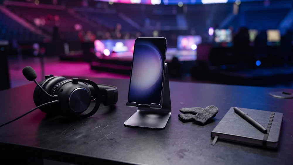 A smartphone in a stand, gaming headset, finger sleeves and a notepad on a tournament desk in an esports arena