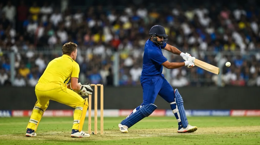 A batsman in blue driving a white cricket ball under floodlights at night while a wicketkeeper in yellow crouches behind the stumps