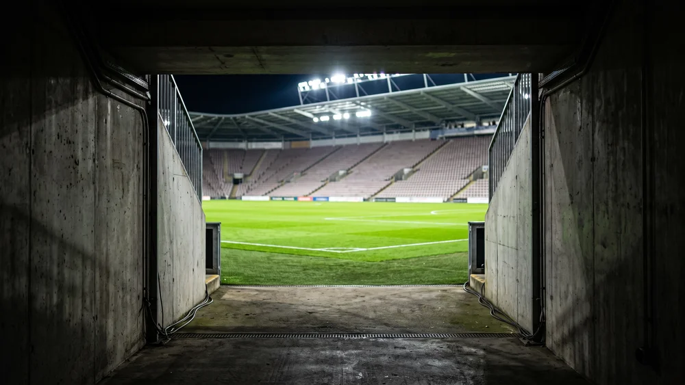 The view out of a dark stadium players tunnel towards a floodlit pitch and empty stand