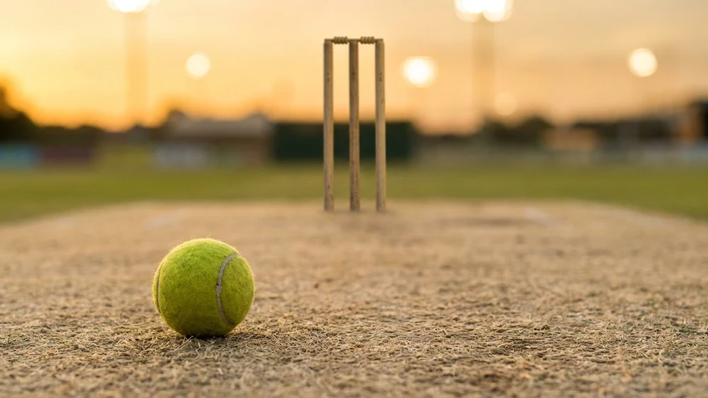 A tennis ball resting on a hard cricket pitch with stumps and bails in the background at dusk