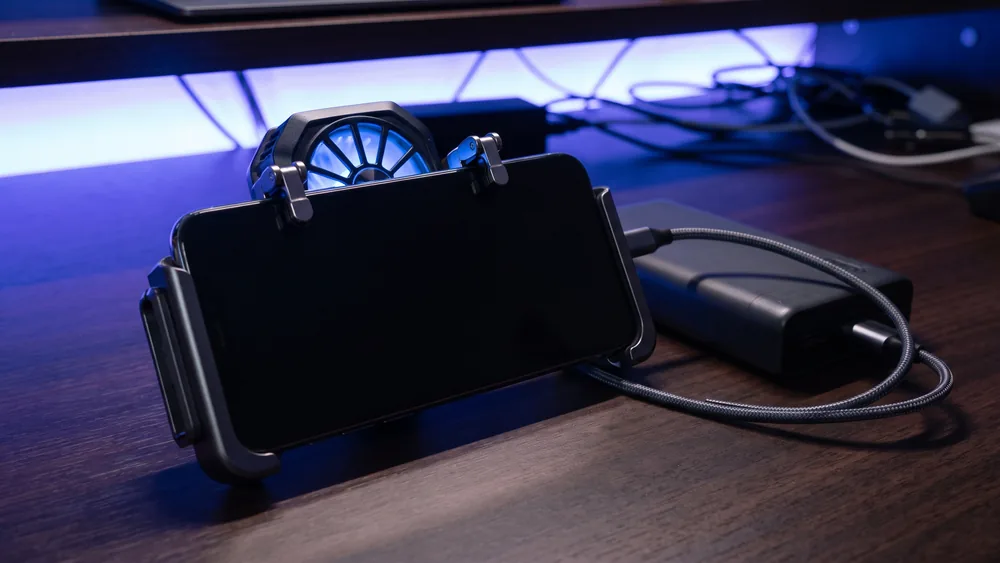 A smartphone in a gaming clamp with a clip-on cooling fan, shoulder trigger buttons and a power bank on a dark desk