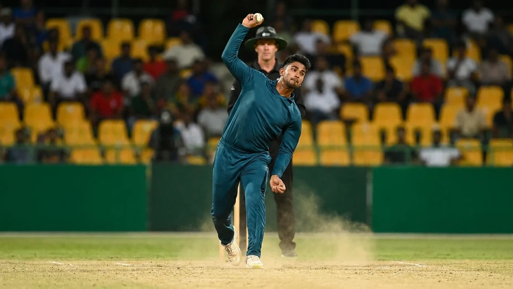 A spin bowler in his delivery stride during a day-night cricket match, dust rising from a dry pitch