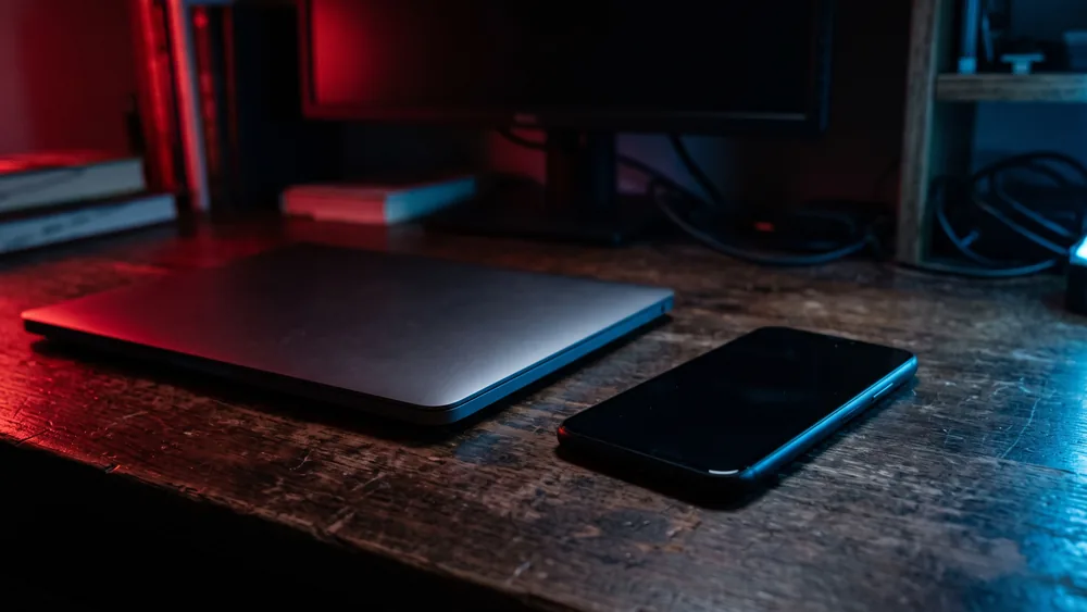 A smartphone with a blank screen next to a closed laptop on a desk lit in red and blue