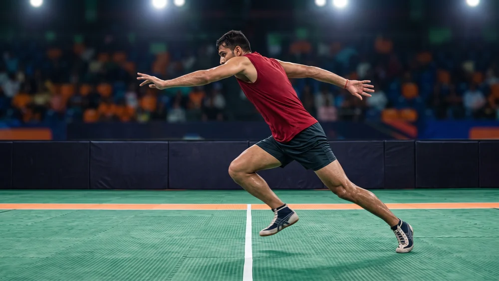 A kabaddi raider in a red jersey and dark shorts crossing the white midline of a green mat during an indoor league match