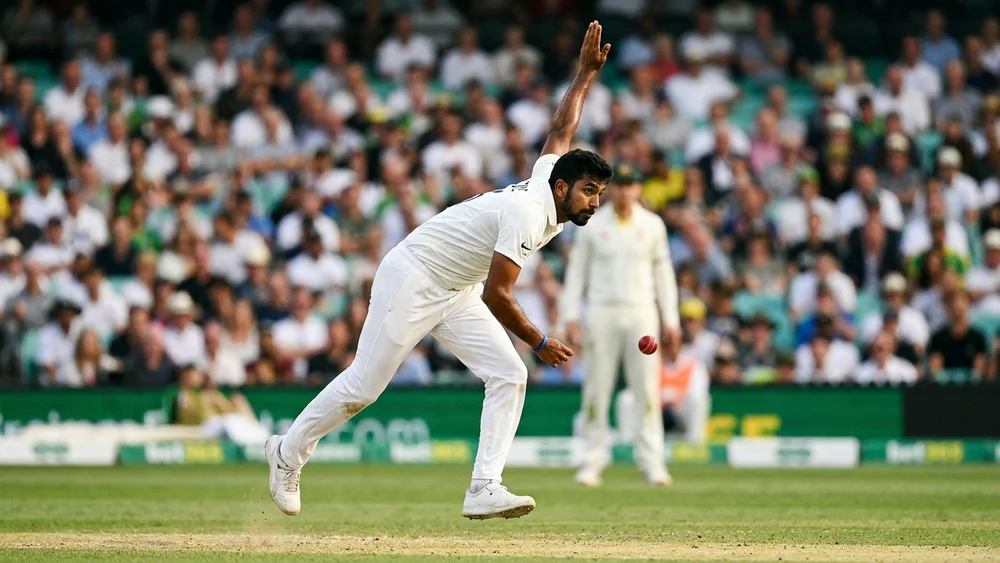 A bowler in white cricket clothing follows through in his delivery stride with a red ball in the air, packed stadium stands blurred behind him