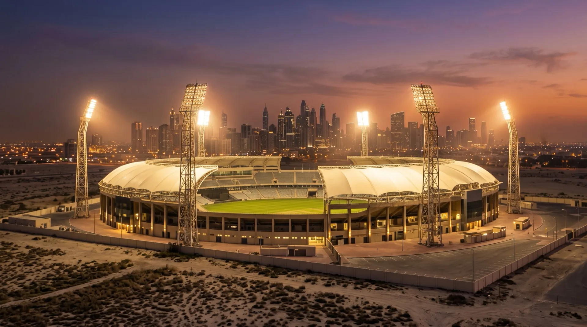 A floodlit cricket stadium at dusk in a Gulf desert city with high-rise towers behind it