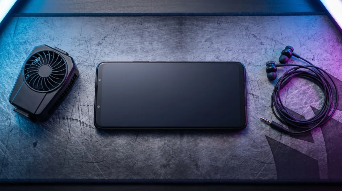 A gaming smartphone lying on a desk mat beside a clip-on cooling fan and earphones under blue and magenta light