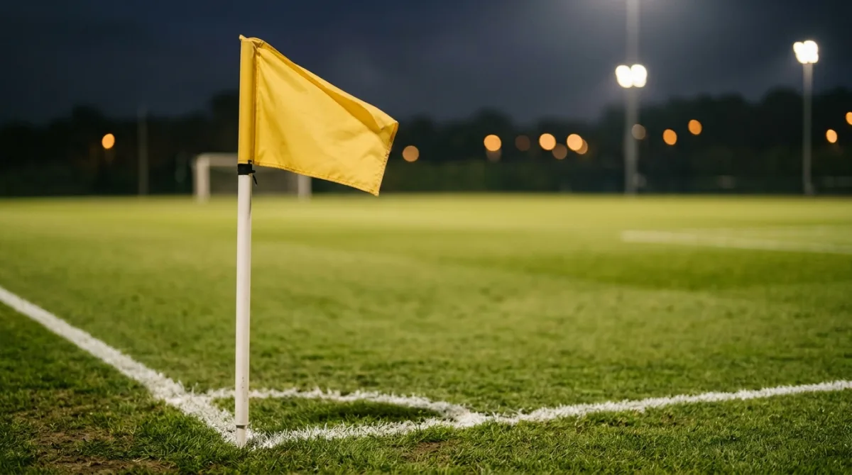 A yellow corner flag on a floodlit football pitch at dusk