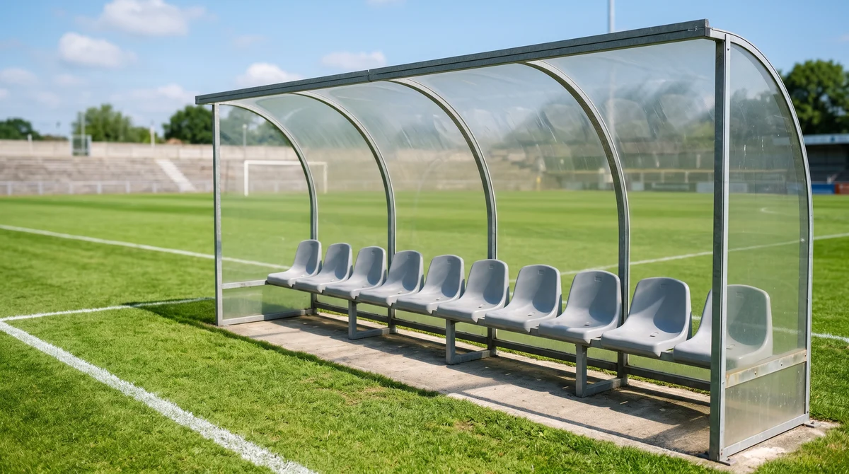 An empty team dugout of plain seats beside a sunlit football pitch
