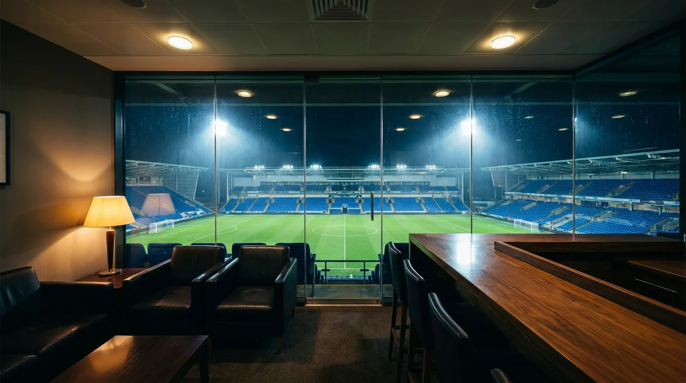 View from an empty executive hospitality box through glass onto a floodlit football pitch and empty stands at night