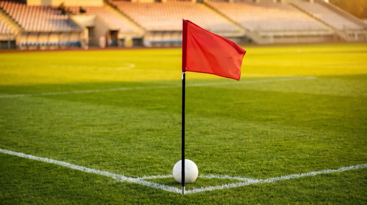 A corner flag and a white football on the corner arc of a sunlit football pitch