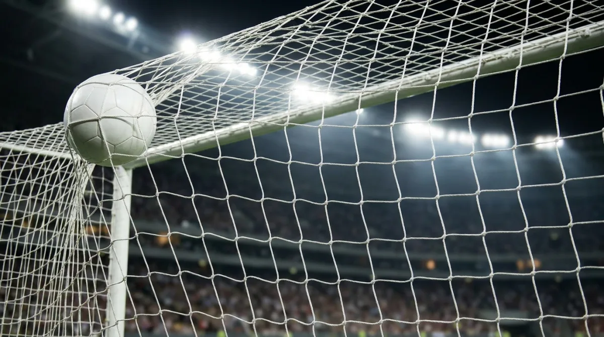 A football in the top corner of the goal net seen from behind the goal under stadium floodlights