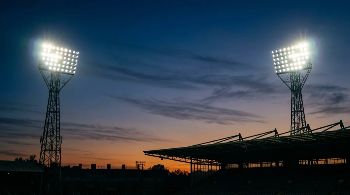 Two stadium floodlight pylons lit against a deep blue dusk sky above a silhouetted stand