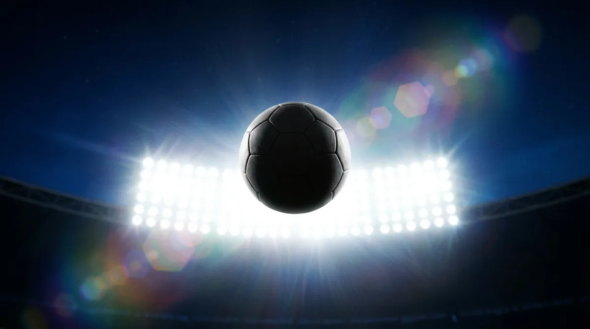 A football in mid-air silhouetted against a bank of stadium floodlights at night