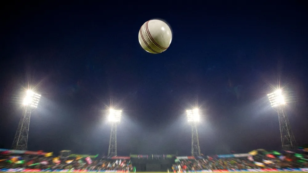 A white cricket ball high against floodlights above an out-of-focus stadium crowd