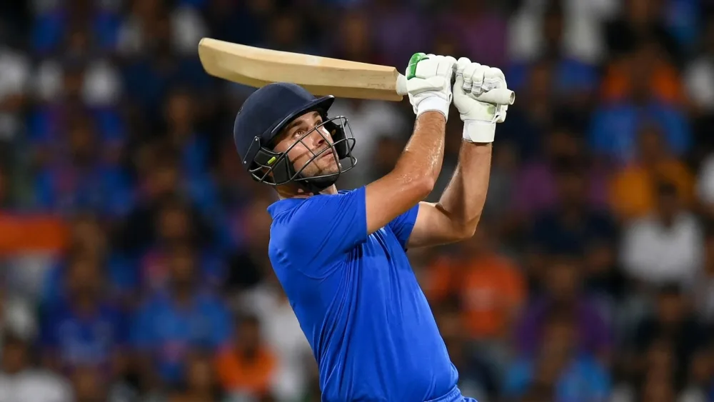 A batsman in plain blue kit swinging through a lofted shot under floodlights