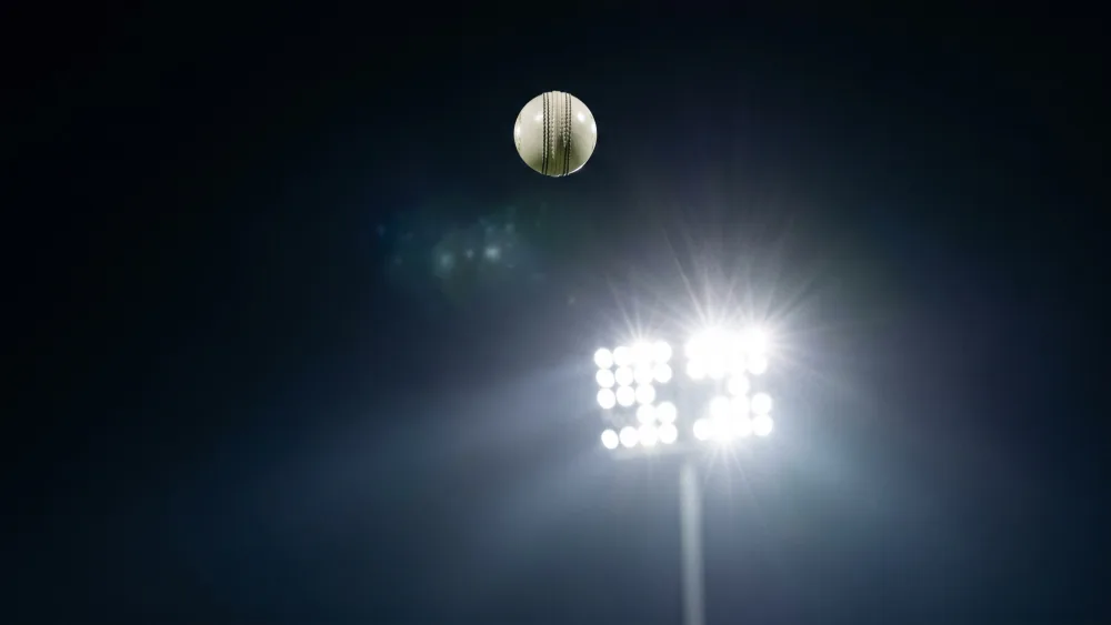 A white cricket ball high in the night sky above a bank of stadium floodlights