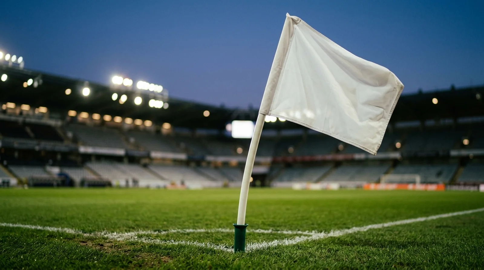 A plain white corner flag on the grass of a floodlit football stadium at dusk
