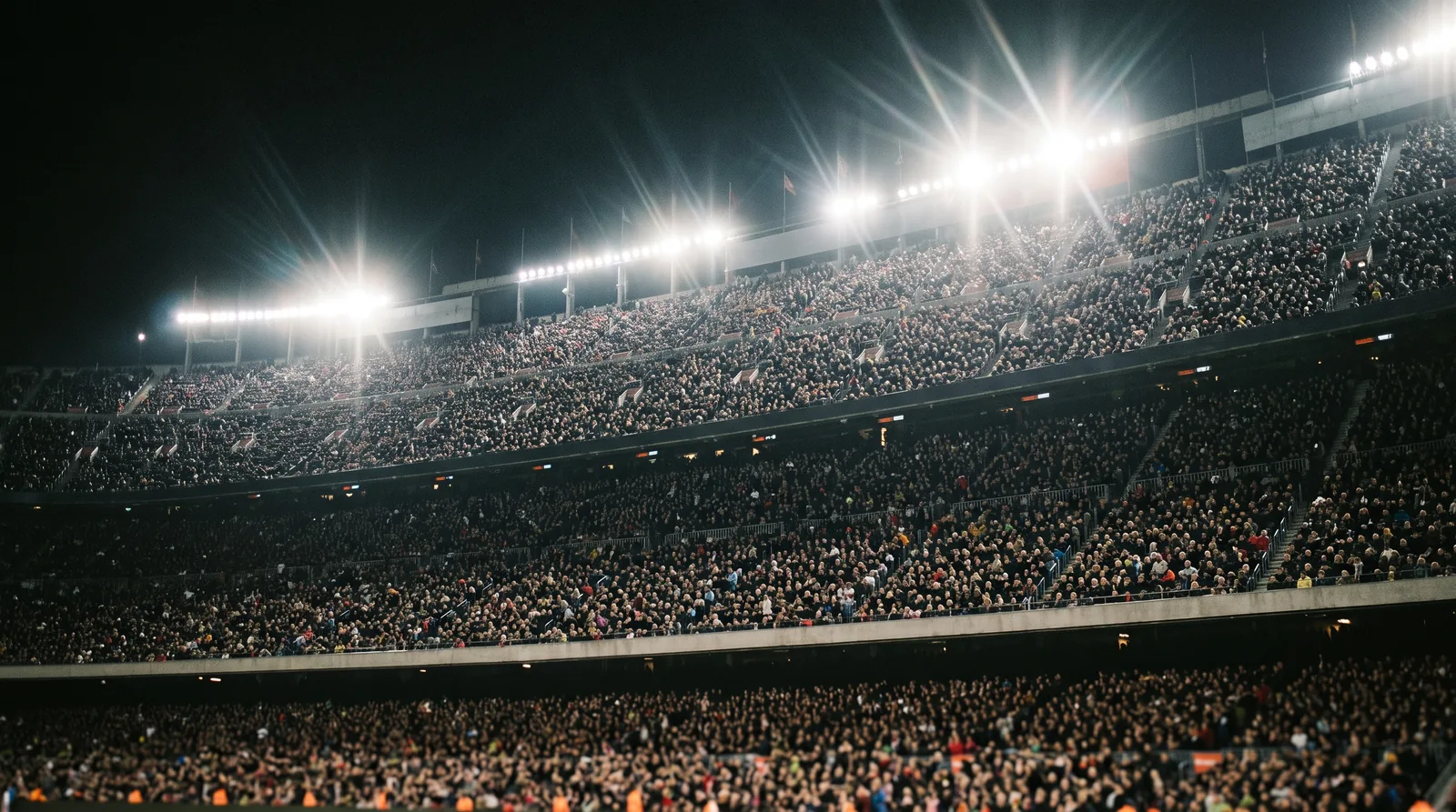 A packed stadium stand under floodlights at night, seen from pitch level