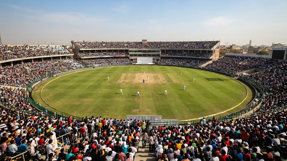A cricket ground in daylight seen from the stands, with players spread across a green outfield and a packed crowd behind them