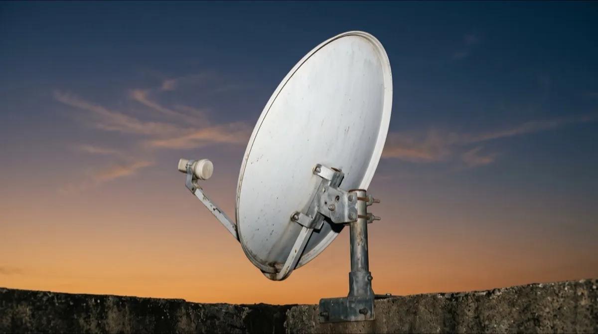 A satellite television dish mounted on a rooftop parapet against a twilight sky