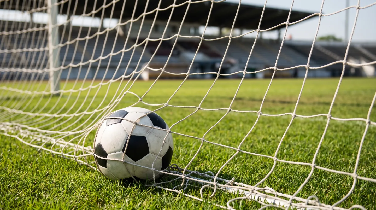 A football resting in the back of a goal net on green grass with empty stadium stands blurred behind