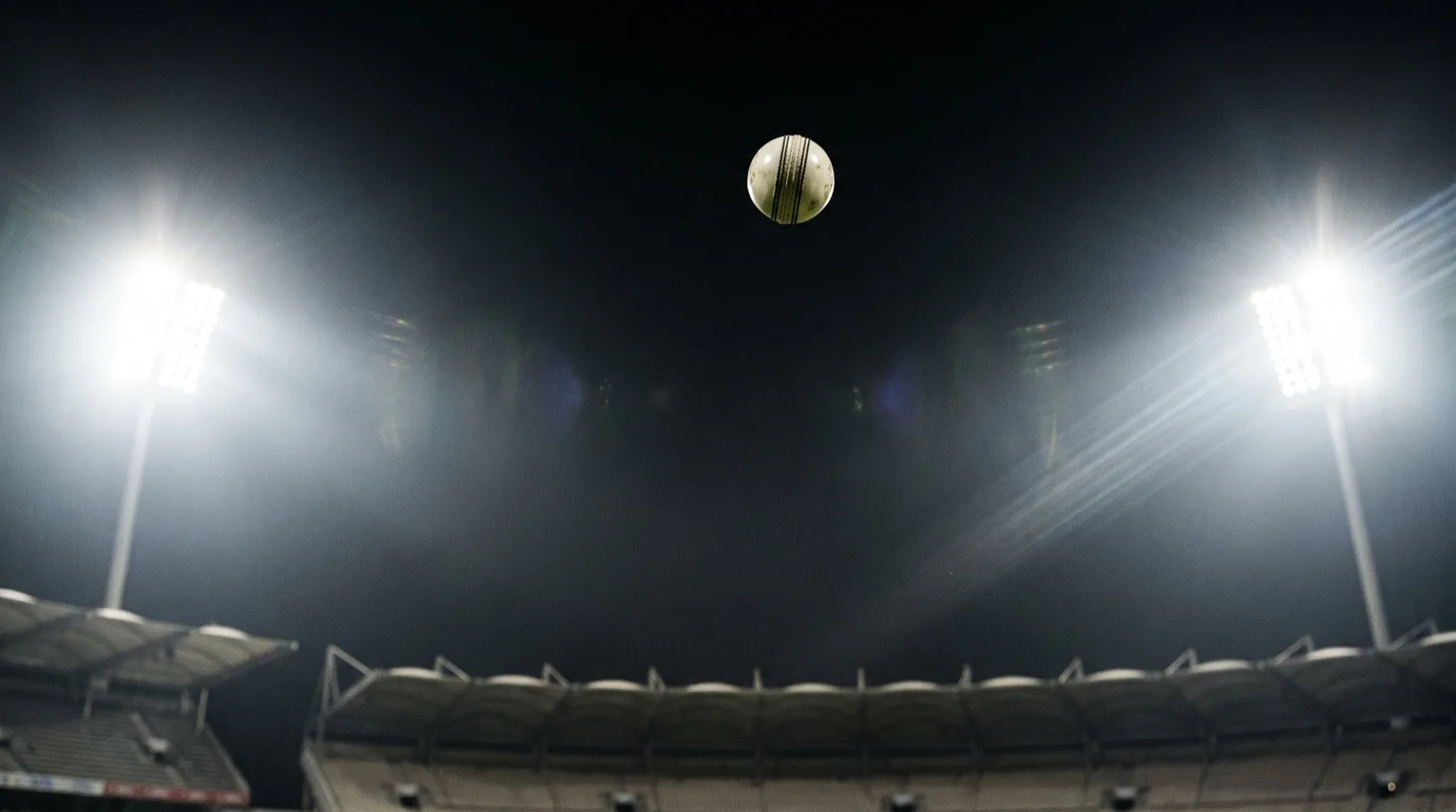 A white cricket ball high in the night sky above a floodlit stadium