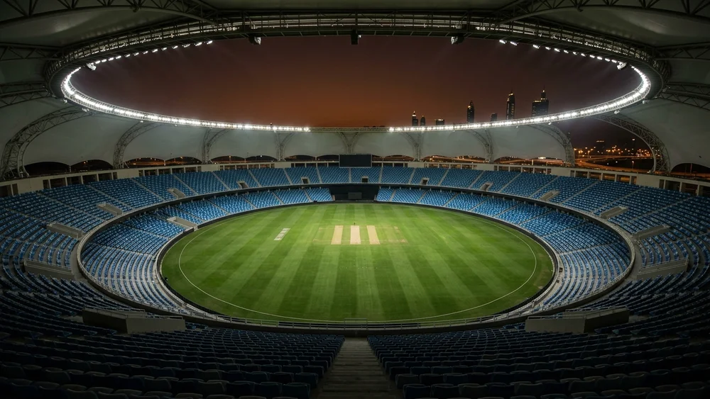 An empty floodlit cricket stadium in a Gulf city at night, seen from the upper tier