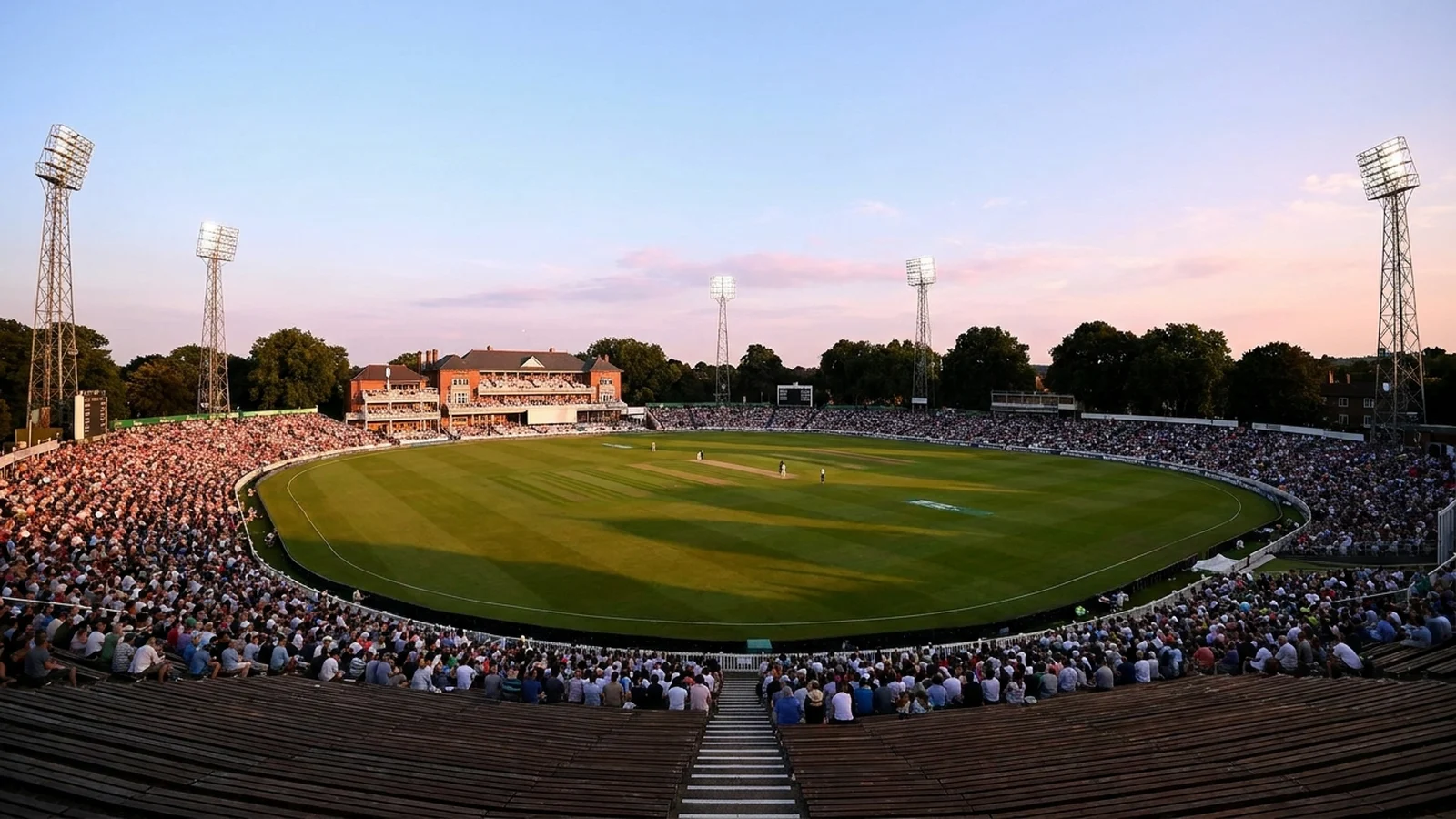 A packed English cricket ground under floodlights at dusk, seen from high in the stands