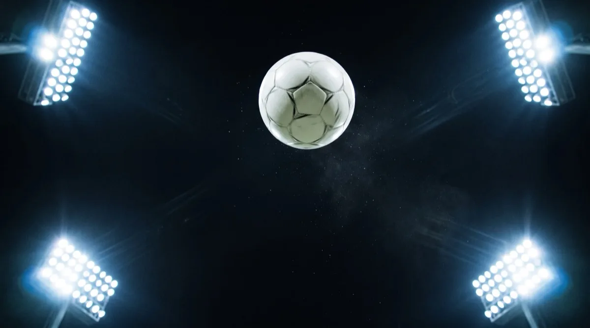 A white football high in the air above a stadium pitch at night, lit by floodlights