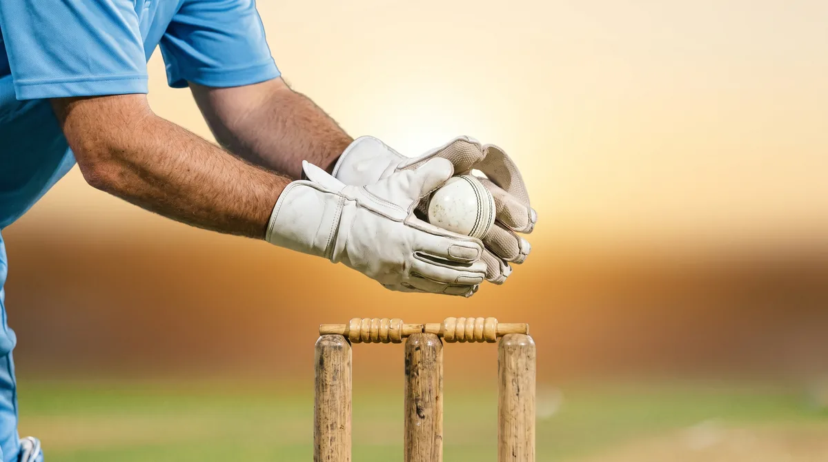 A wicketkeeper gathers a white ball in his gloves behind the stumps