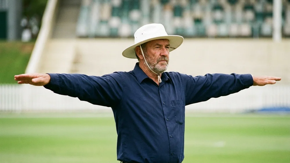 A cricket umpire in a navy shirt and white sun hat signalling a wide with both arms stretched out sideways