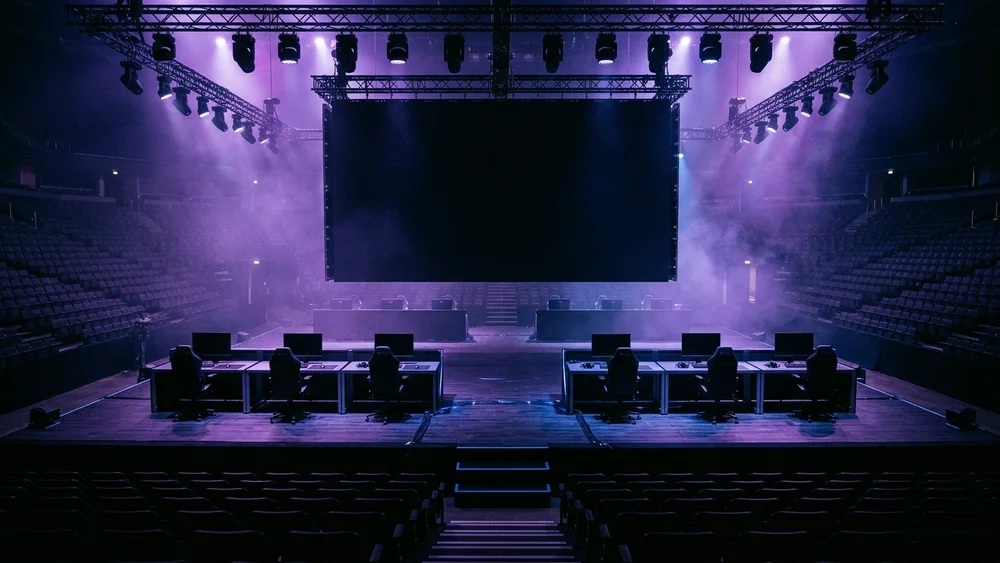 An empty esports arena stage with gaming desks under purple lights before a final