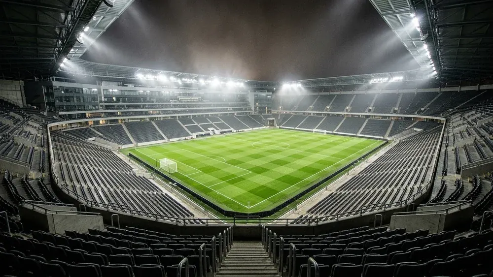 An empty modern football stadium at night under floodlights, with grey seats in every stand and a striped green pitch