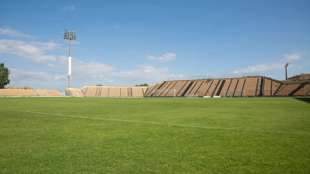 An empty football stadium with a green pitch and rows of orange seats under a blue sky