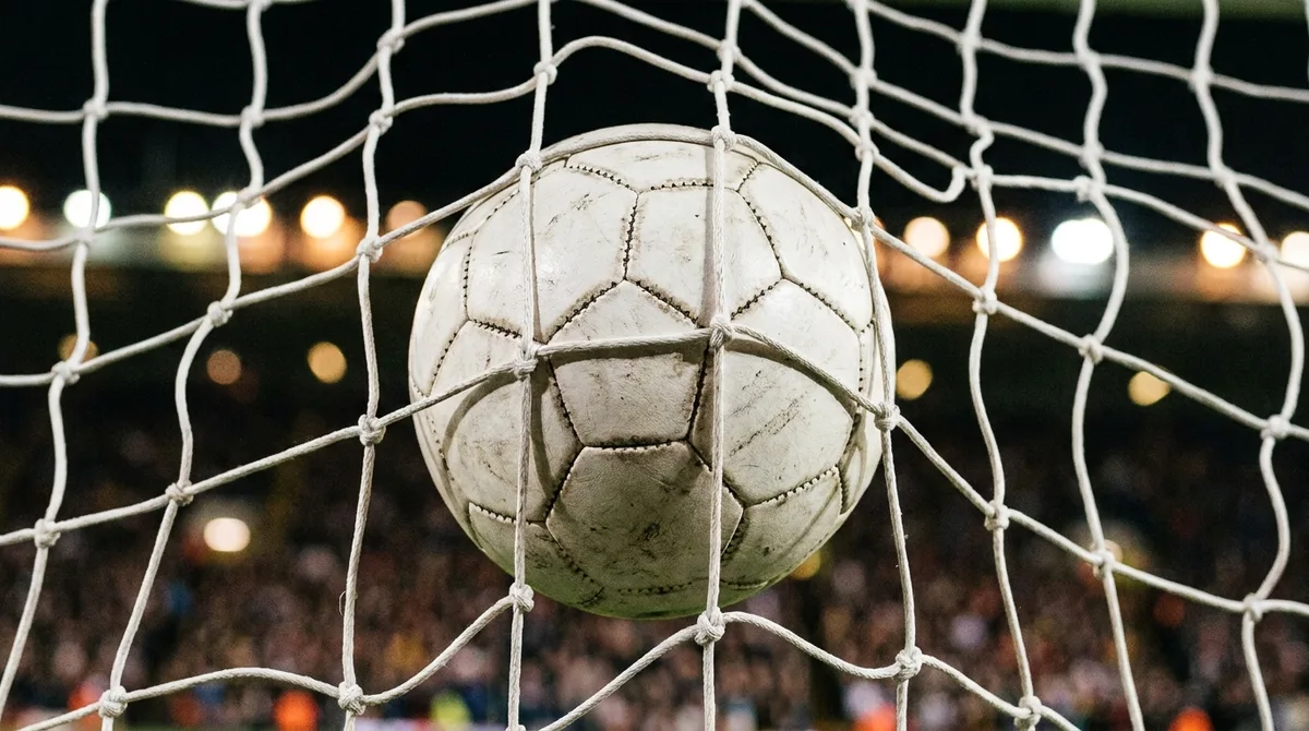 A football bulging the back of the net under stadium floodlights