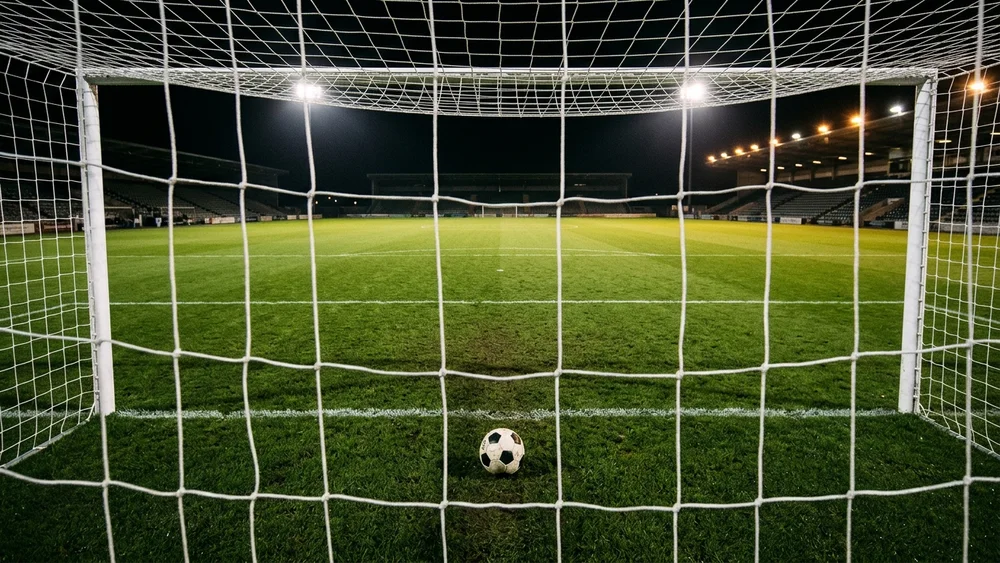 A football resting on the grass just inside the goal, seen through the net, with an empty floodlit pitch beyond