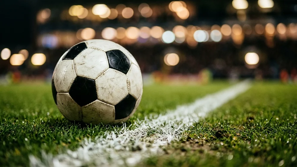 A worn white football on the grass next to a white pitch line under floodlights