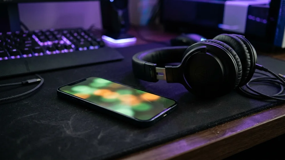 A smartphone lying on a gaming desk beside a pair of black headphones under purple light