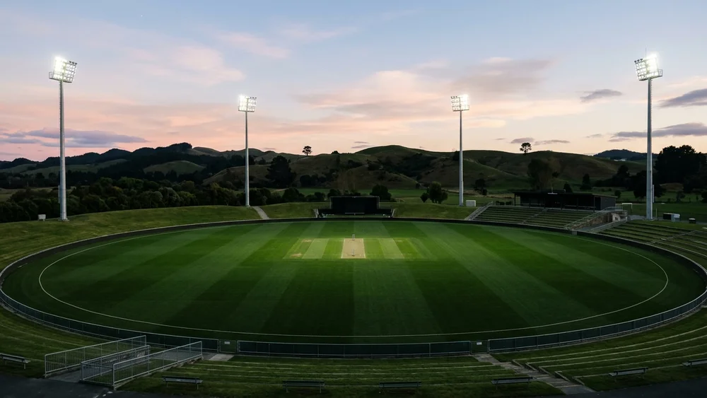 An empty floodlit cricket ground at dusk with green hills behind