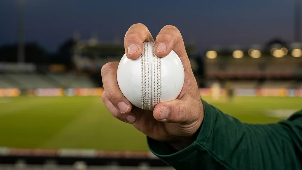 A spinner in a green one-day shirt grips a white ball across the seam at a floodlit ground